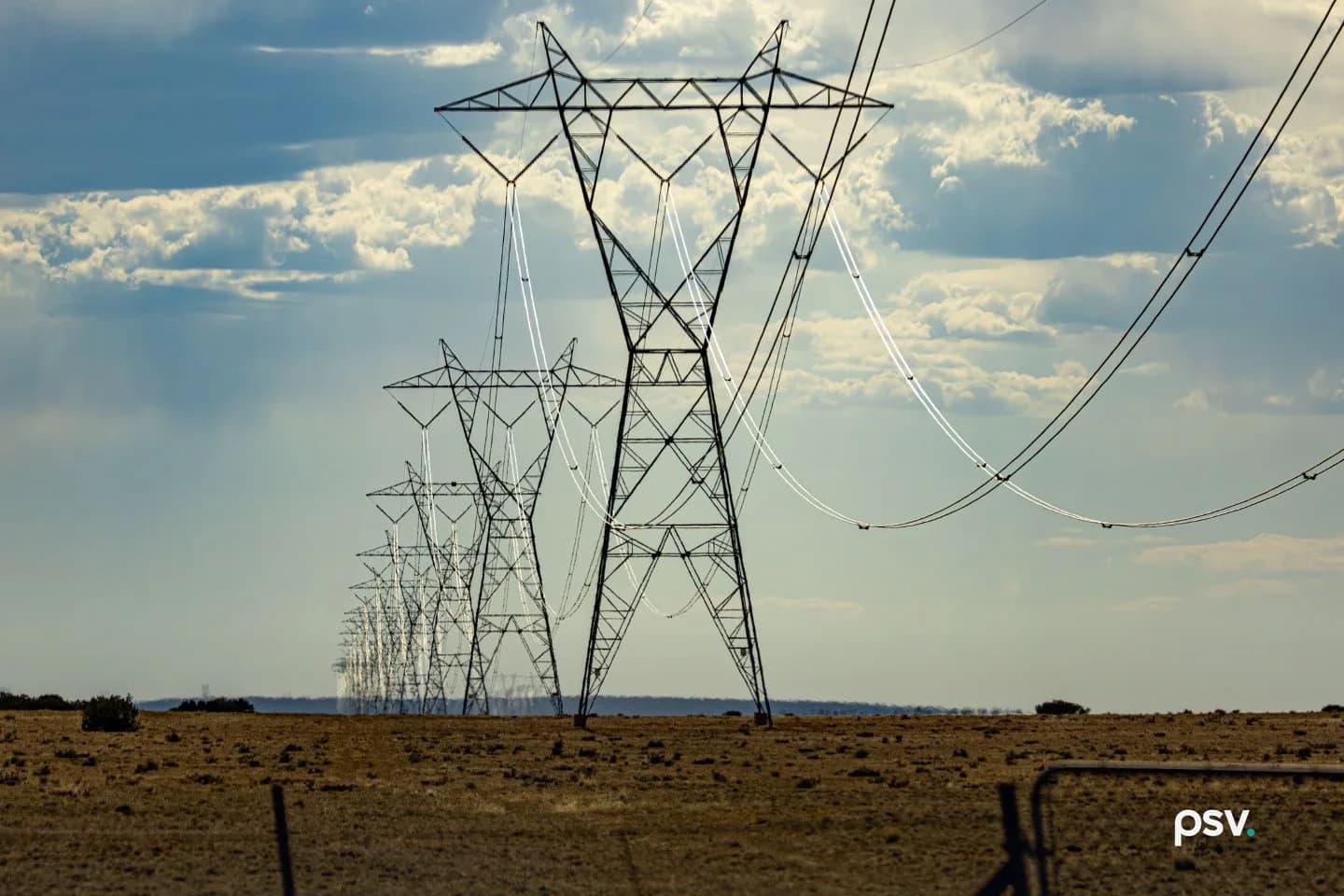 A row of tall steel lattice transmission towers carrying high-voltage lines across a flat, dry brown field, receding to the horizon under a blue sky with scattered white clouds, a wire fence and a metal gate in the foreground at right. A row of tall steel lattice transmission towers carrying high-voltage lines across a flat, dry brown field, receding to the horizon under a blue sky with scattered white clouds, a wire fence and a metal gate in the foreground at right.