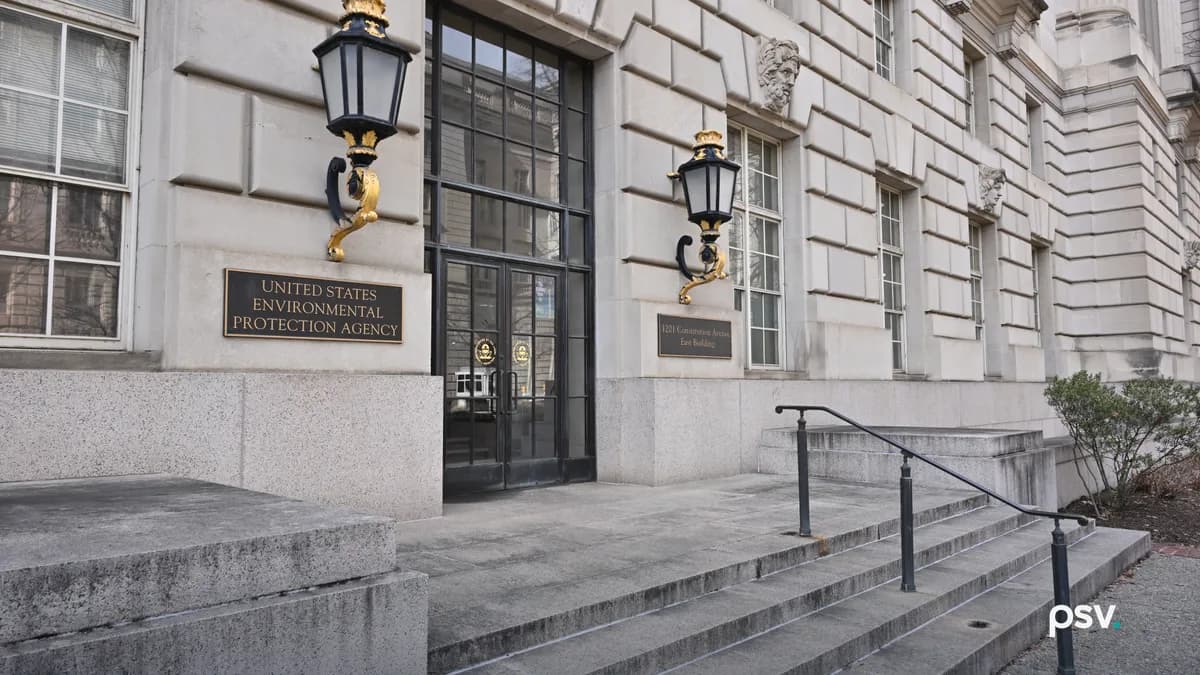 A daylight street-level view of the limestone entrance to the east building of the United States Environmental Protection Agency at 1201 Constitution Avenue NW in Washington, DC, with a brass plaque reading UNITED STATES ENVIRONMENTAL PROTECTION AGENCY beside tall black-framed glass doors, two gilded lanterns on the wall, and empty granite steps in the foreground.