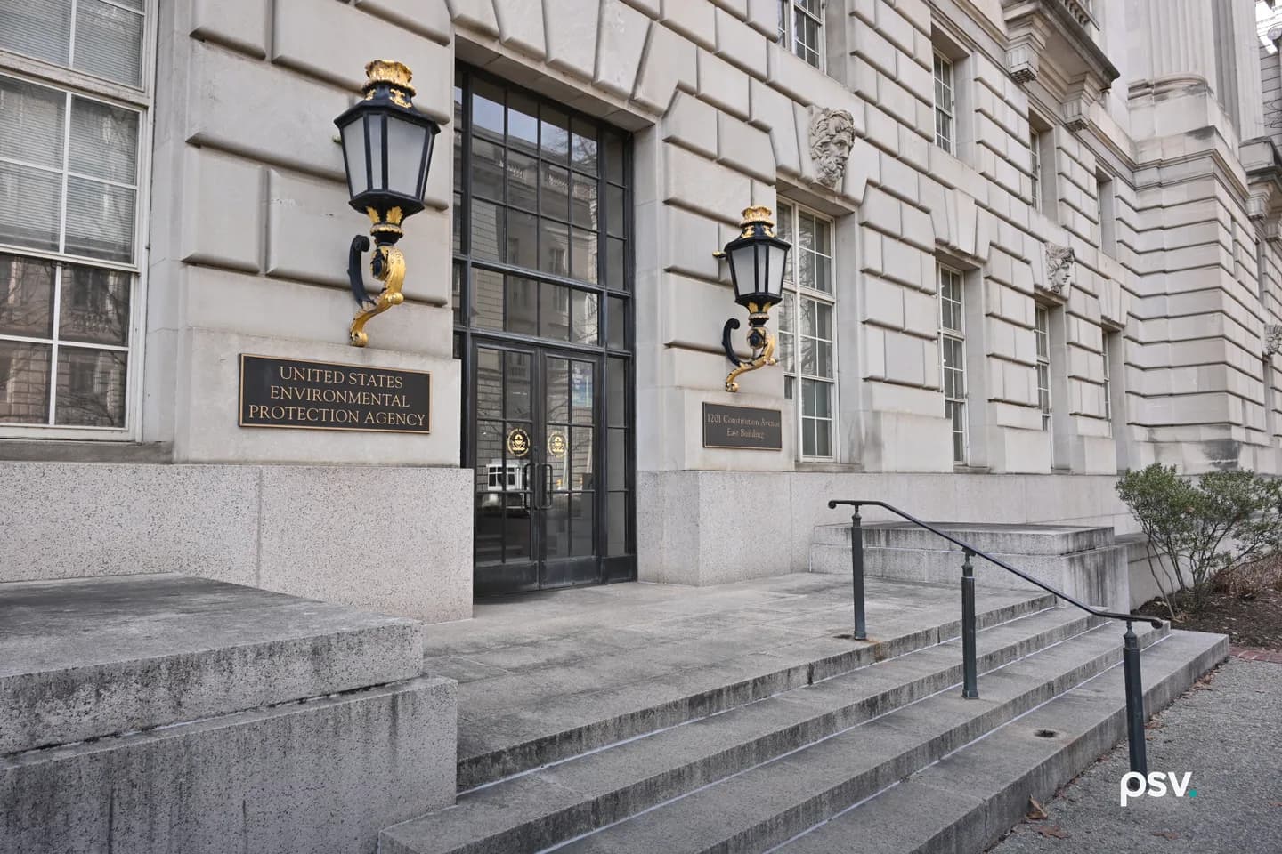 A daylight street-level view of the limestone entrance to the east building of the United States Environmental Protection Agency at 1201 Constitution Avenue NW in Washington, DC, with a brass plaque reading UNITED STATES ENVIRONMENTAL PROTECTION AGENCY beside tall black-framed glass doors, two gilded lanterns on the wall, and empty granite steps in the foreground. A daylight street-level view of the limestone entrance to the east building of the United States Environmental Protection Agency at 1201 Constitution Avenue NW in Washington, DC, with a brass plaque reading UNITED STATES ENVIRONMENTAL PROTECTION AGENCY beside tall black-framed glass doors, two gilded lanterns on the wall, and empty granite steps in the foreground.