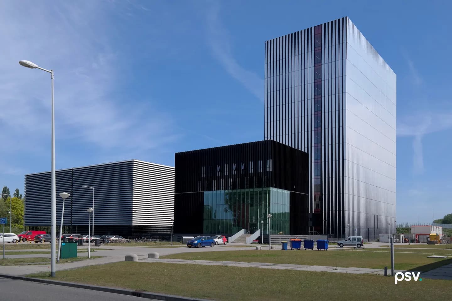 A low black-and-white horizontally louvered data center building on the left, a black-clad cube with a glass entrance in the middle, and a tall slender glass-and-metal tower on the right, seen from a grassy street corner under a blue sky with thin clouds. A low black-and-white horizontally louvered data center building on the left, a black-clad cube with a glass entrance in the middle, and a tall slender glass-and-metal tower on the right, seen from a grassy street corner under a blue sky with thin clouds.