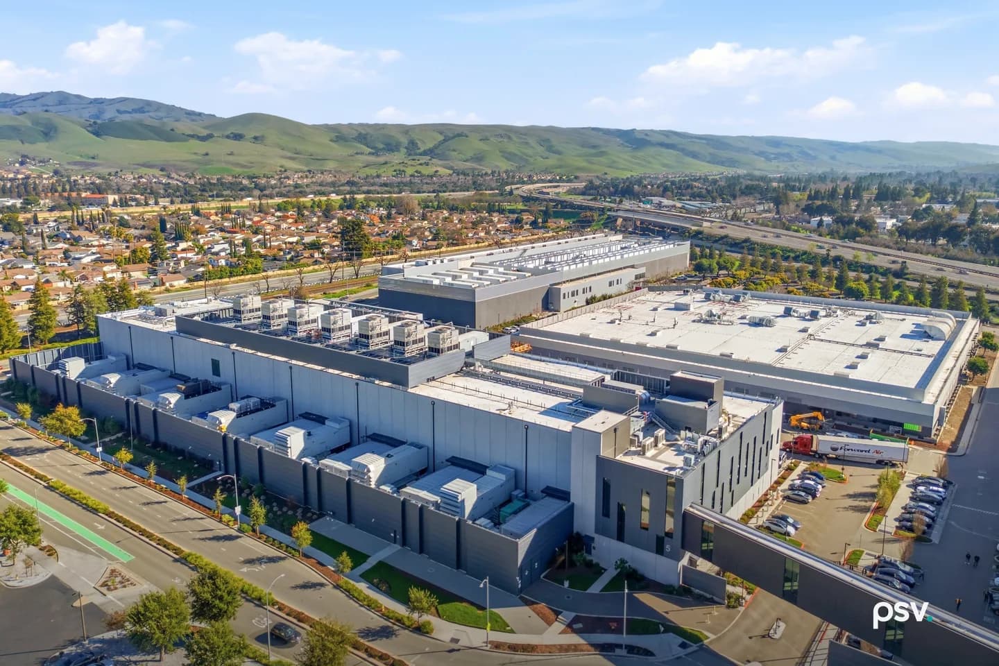 An aerial daylight photograph of a large Equinix data center campus: a long grey windowless building with rows of white cooling units along its roof and side yard fronts a landscaped street with a green bike lane, a second flat-roofed building and a taller rooftop-equipment block sit behind it, a truck and parked cars fill a lot at right, and suburban houses, a freeway and green hills stretch to the horizon under a blue sky with scattered clouds. An aerial daylight photograph of a large Equinix data center campus: a long grey windowless building with rows of white cooling units along its roof and side yard fronts a landscaped street with a green bike lane, a second flat-roofed building and a taller rooftop-equipment block sit behind it, a truck and parked cars fill a lot at right, and suburban houses, a freeway and green hills stretch to the horizon under a blue sky with scattered clouds.