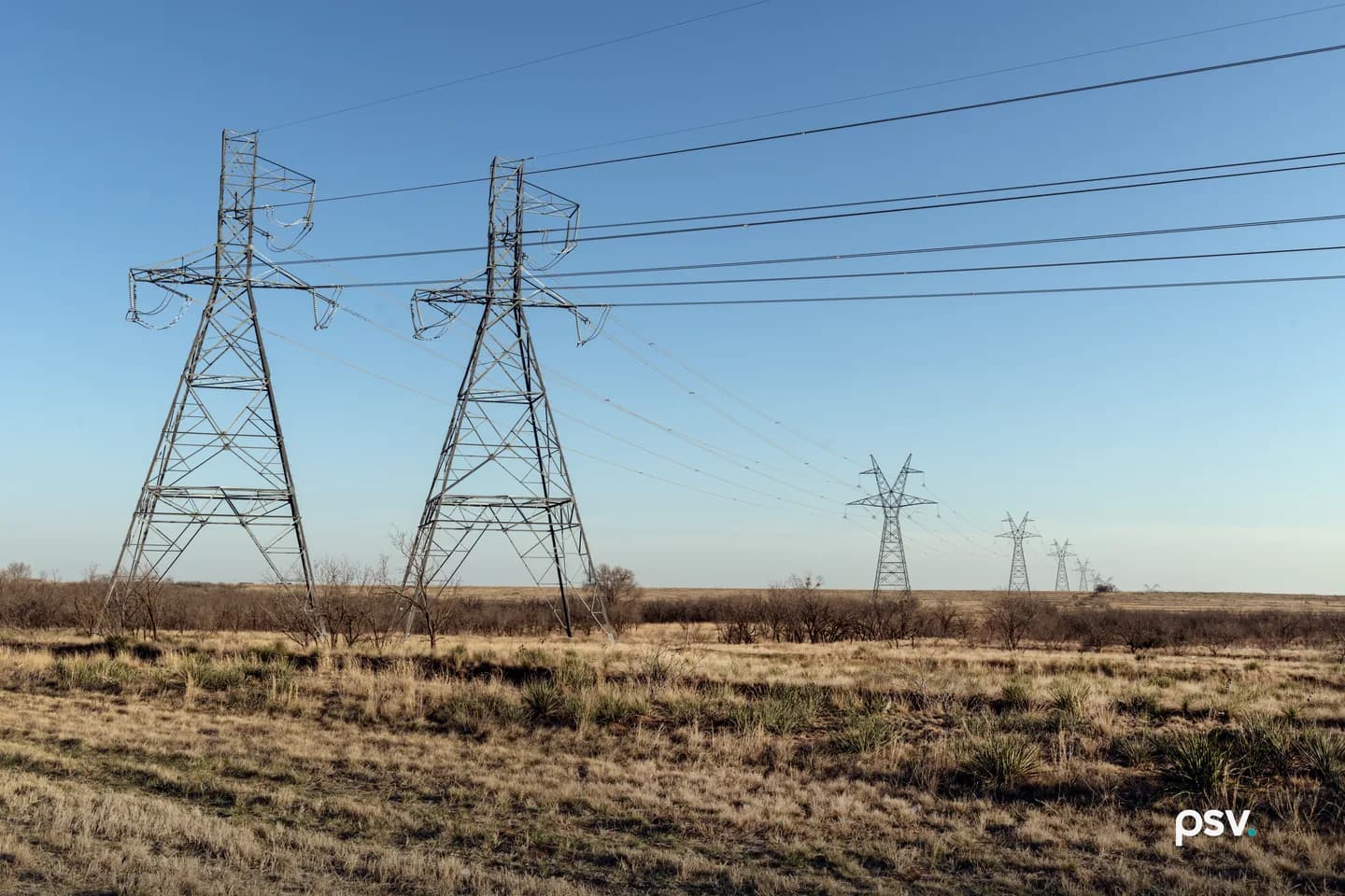 A daylight view across flat West Texas scrubland of two tall steel lattice transmission towers in the left foreground carrying multiple conductor lines that sweep to the right, with a receding line of smaller towers marching toward a low horizon under a clear pale blue sky, dry gold grass and bare mesquite in the foreground. A daylight view across flat West Texas scrubland of two tall steel lattice transmission towers in the left foreground carrying multiple conductor lines that sweep to the right, with a receding line of smaller towers marching toward a low horizon under a clear pale blue sky, dry gold grass and bare mesquite in the foreground.