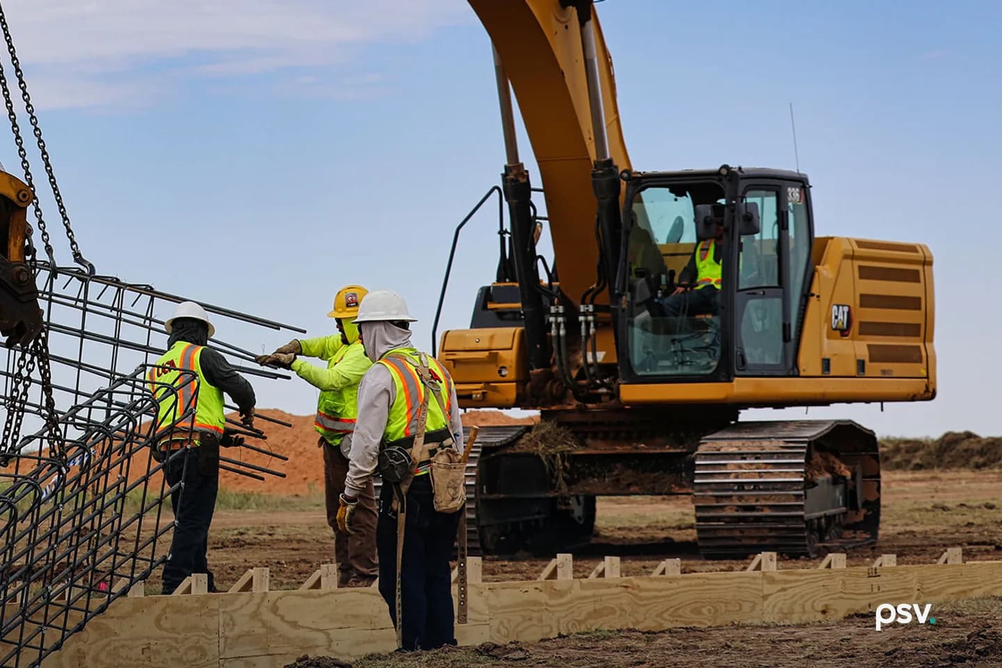 Three workers in high-visibility vests and hard hats guiding a long steel reinforcing cage down into timber formwork on bare prairie ground, one of them steadying a bundle of rebar, with a tracked CAT 336 excavator and its operator behind them and mounds of red excavated soil on the horizon under a pale sky Three workers in high-visibility vests and hard hats guiding a long steel reinforcing cage down into timber formwork on bare prairie ground, one of them steadying a bundle of rebar, with a tracked CAT 336 excavator and its operator behind them and mounds of red excavated soil on the horizon under a pale sky