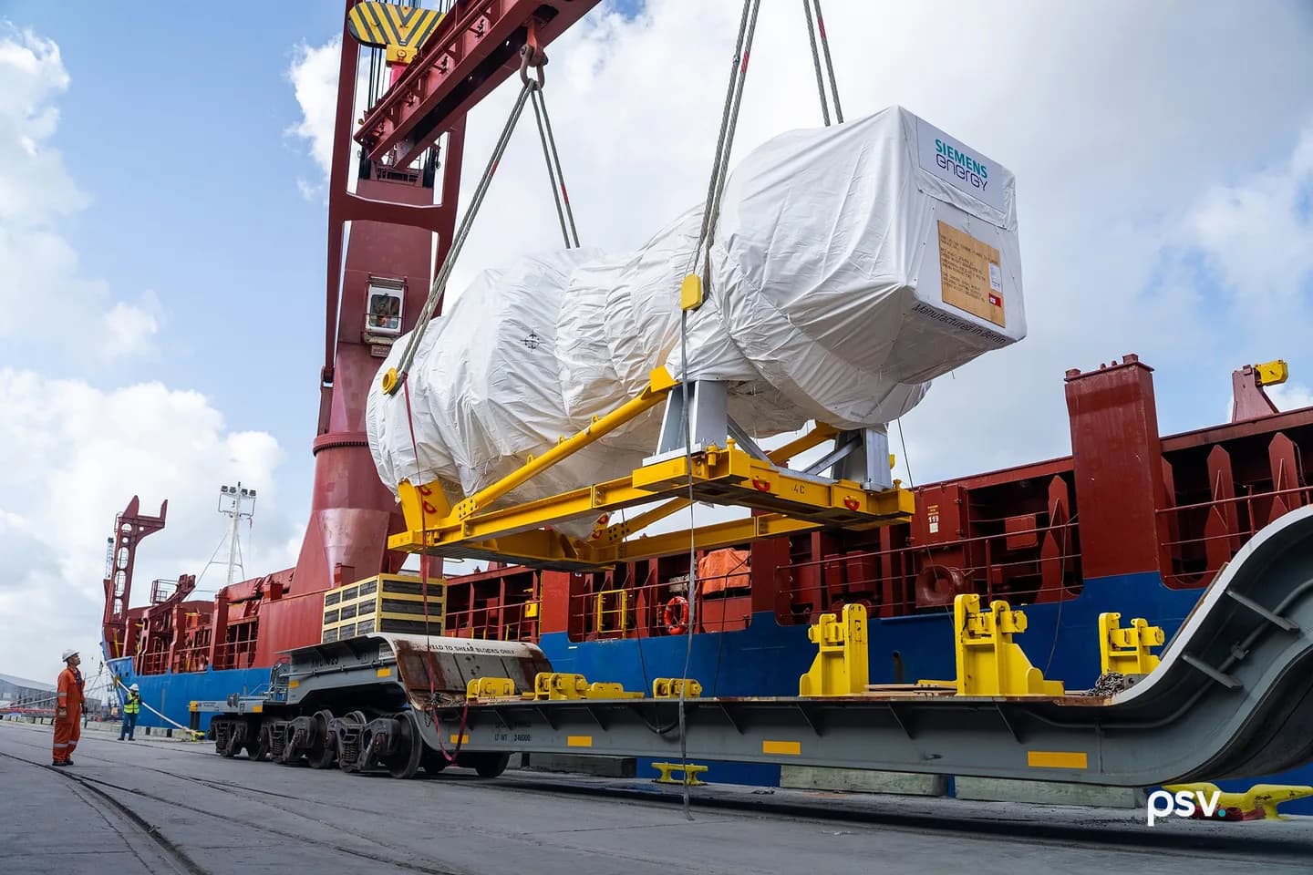 A dockside gantry crane lifting a shrink-wrapped Siemens Energy gas turbine on a yellow lifting frame off the deck of a red and blue heavy-lift ship and down toward a multi-axle rail transporter on the quay, with two workers in orange and yellow high-visibility gear watching from the left under a bright cloudy sky A dockside gantry crane lifting a shrink-wrapped Siemens Energy gas turbine on a yellow lifting frame off the deck of a red and blue heavy-lift ship and down toward a multi-axle rail transporter on the quay, with two workers in orange and yellow high-visibility gear watching from the left under a bright cloudy sky