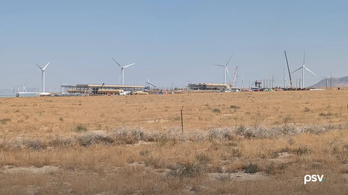 A daylight view across flat, dry high-desert rangeland in Utah toward a geothermal plant under construction on the horizon: long low steel structures and open pipe racks, a large horizontal tank, a silo, scattered stacked materials and site trailers, a yellow crawler crane with its boom raised at right, a line of utility poles running to the right edge, and a row of white wind turbines standing behind and beyond the plant under a pale blue sky, with a barbed wire fence and gold grass in the foreground.