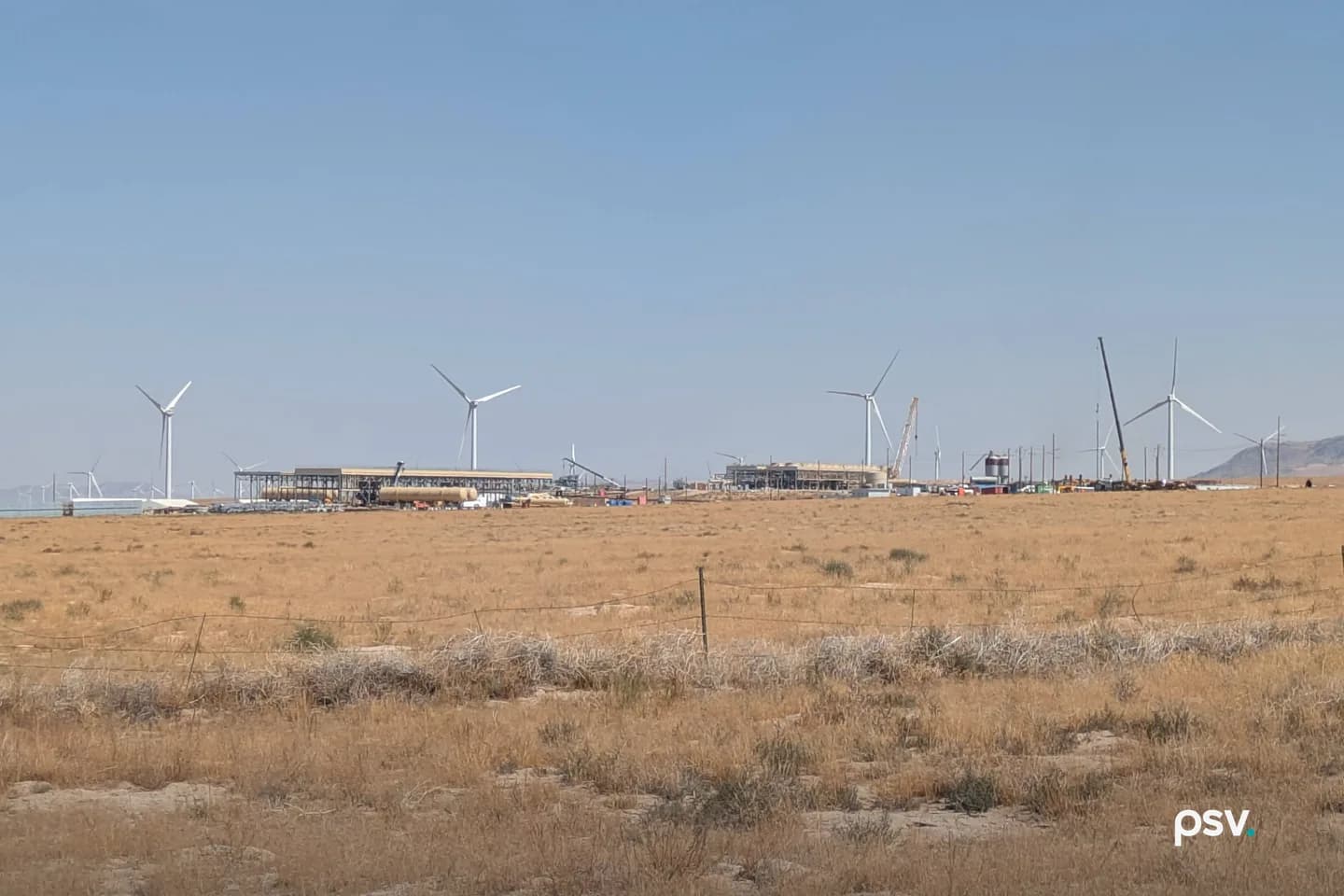 A daylight view across flat, dry high-desert rangeland in Utah toward a geothermal plant under construction on the horizon: long low steel structures and open pipe racks, a large horizontal tank, a silo, scattered stacked materials and site trailers, a yellow crawler crane with its boom raised at right, a line of utility poles running to the right edge, and a row of white wind turbines standing behind and beyond the plant under a pale blue sky, with a barbed wire fence and gold grass in the foreground. A daylight view across flat, dry high-desert rangeland in Utah toward a geothermal plant under construction on the horizon: long low steel structures and open pipe racks, a large horizontal tank, a silo, scattered stacked materials and site trailers, a yellow crawler crane with its boom raised at right, a line of utility poles running to the right edge, and a row of white wind turbines standing behind and beyond the plant under a pale blue sky, with a barbed wire fence and gold grass in the foreground.