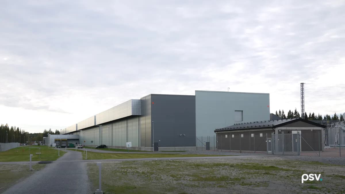 A daylight photograph under a pale overcast sky of a long, windowless hyperscale data center building clad in grey, silver and pale green metal panels, stretching away to the left toward a loading dock and a line of conifers, with a smaller brown single-storey electrical building behind a chain-link fence and gate at right, a lattice transmission mast and part of a substation at the far right edge, a gravel path and low bollard lights crossing a patchy grass and gravel lot in the foreground, and two parked cars beside the building.