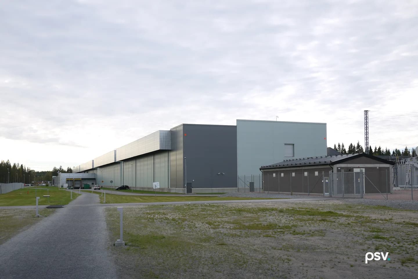 A daylight photograph under a pale overcast sky of a long, windowless hyperscale data center building clad in grey, silver and pale green metal panels, stretching away to the left toward a loading dock and a line of conifers, with a smaller brown single-storey electrical building behind a chain-link fence and gate at right, a lattice transmission mast and part of a substation at the far right edge, a gravel path and low bollard lights crossing a patchy grass and gravel lot in the foreground, and two parked cars beside the building. A daylight photograph under a pale overcast sky of a long, windowless hyperscale data center building clad in grey, silver and pale green metal panels, stretching away to the left toward a loading dock and a line of conifers, with a smaller brown single-storey electrical building behind a chain-link fence and gate at right, a lattice transmission mast and part of a substation at the far right edge, a gravel path and low bollard lights crossing a patchy grass and gravel lot in the foreground, and two parked cars beside the building.
