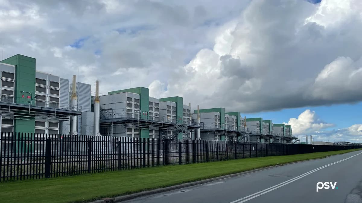 A row of large grey and green industrial enclosures with louvered panels and tall silver exhaust stacks, linked by steel walkways and stairs, standing behind a black steel palisade fence beside a strip of mown grass and an empty two-lane road, under heavy white and grey clouds with a patch of blue sky at the right.