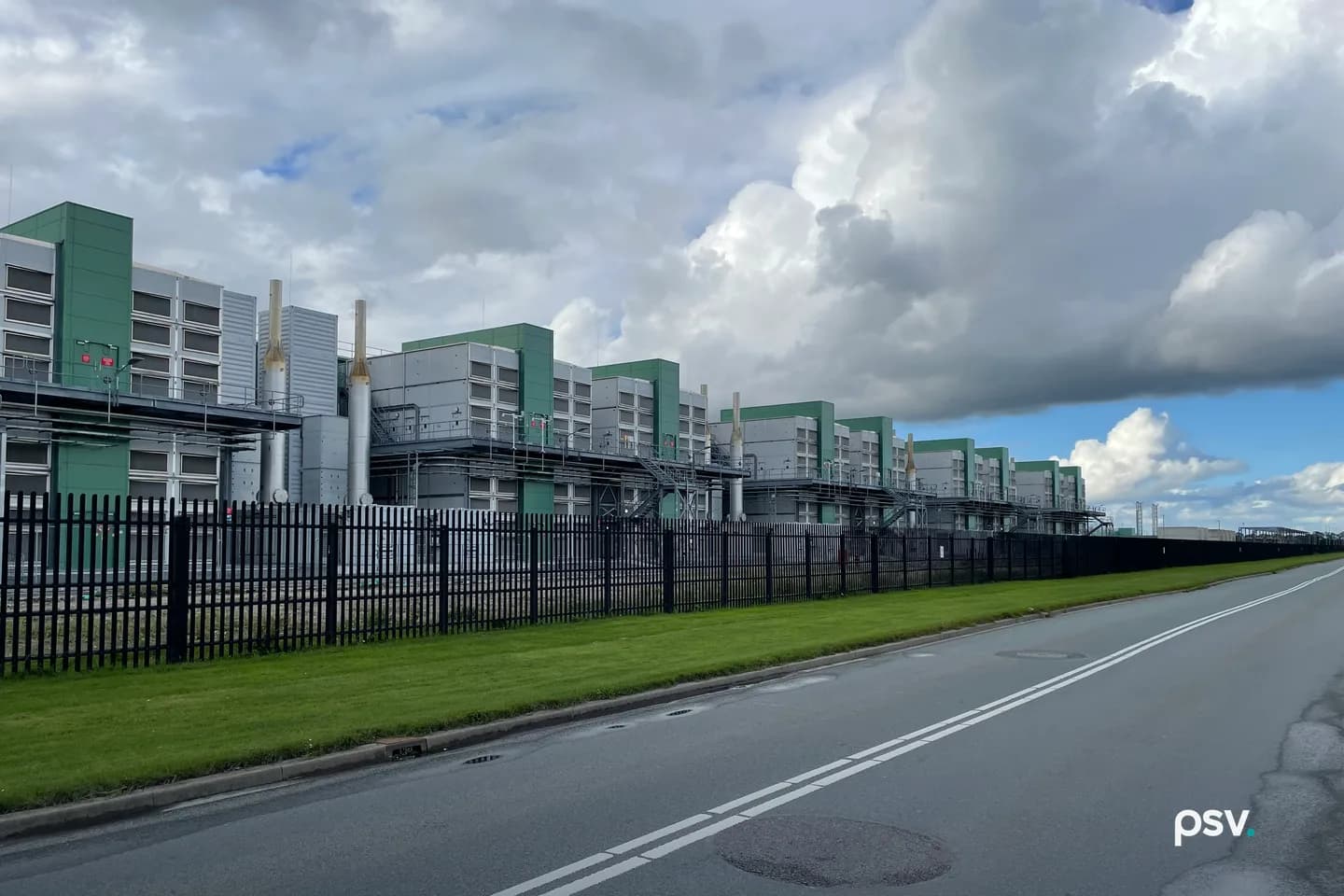 A row of large grey and green industrial enclosures with louvered panels and tall silver exhaust stacks, linked by steel walkways and stairs, standing behind a black steel palisade fence beside a strip of mown grass and an empty two-lane road, under heavy white and grey clouds with a patch of blue sky at the right. A row of large grey and green industrial enclosures with louvered panels and tall silver exhaust stacks, linked by steel walkways and stairs, standing behind a black steel palisade fence beside a strip of mown grass and an empty two-lane road, under heavy white and grey clouds with a patch of blue sky at the right.