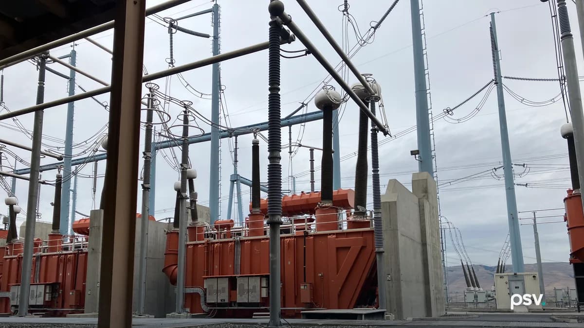 A daylight view inside the yard of a high voltage converter station: two large orange oil-filled converter transformers side by side behind grey concrete blast walls, each topped with tall ribbed black and grey bushings rising into a steel gantry strung with conductors and disconnect switches, pale blue steel supports and lattice towers across the middle ground, transmission poles and overhead lines running to the right toward low brown hills, and a small beige control cabinet building at right under an overcast sky.