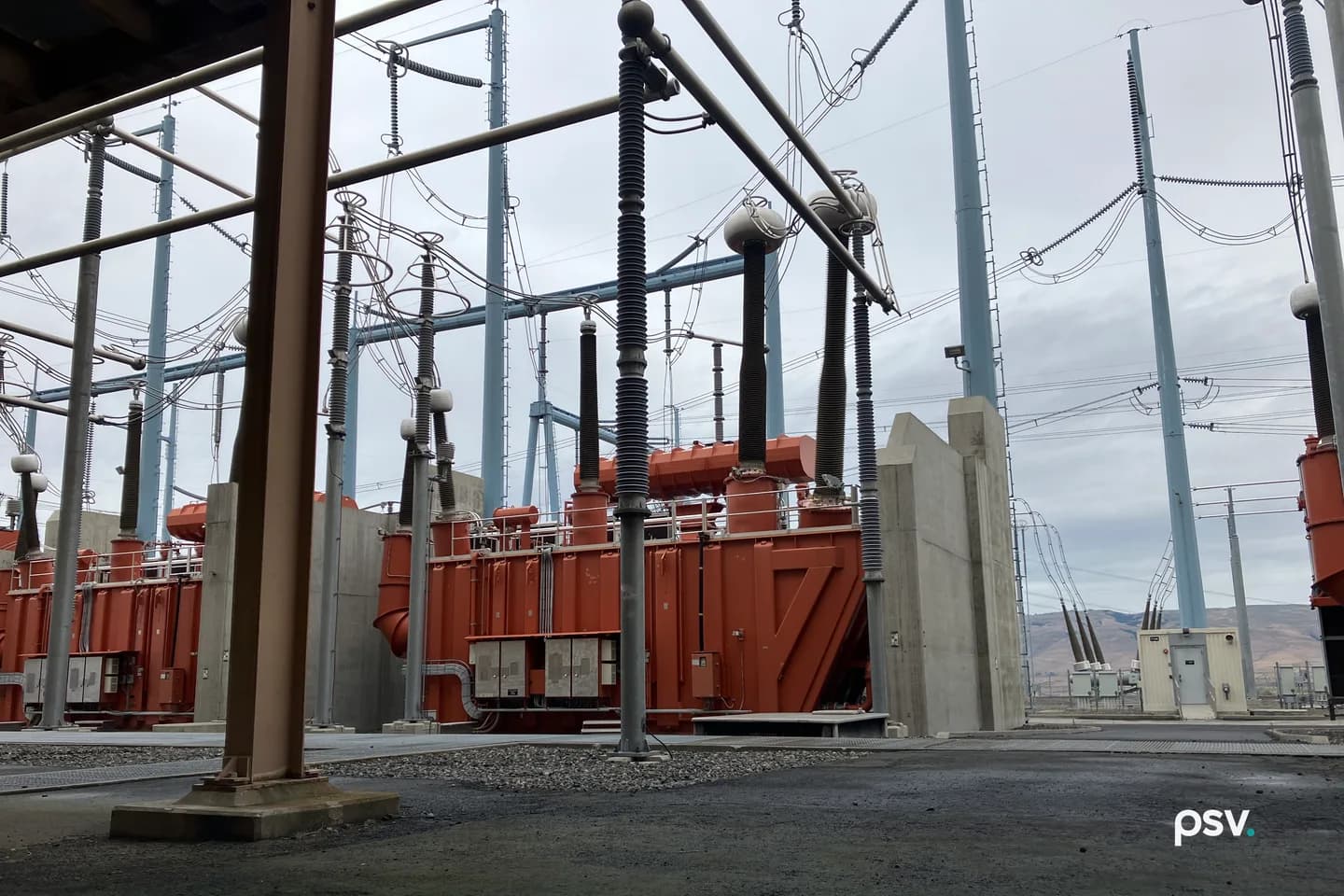 A daylight view inside the yard of a high voltage converter station: two large orange oil-filled converter transformers side by side behind grey concrete blast walls, each topped with tall ribbed black and grey bushings rising into a steel gantry strung with conductors and disconnect switches, pale blue steel supports and lattice towers across the middle ground, transmission poles and overhead lines running to the right toward low brown hills, and a small beige control cabinet building at right under an overcast sky. A daylight view inside the yard of a high voltage converter station: two large orange oil-filled converter transformers side by side behind grey concrete blast walls, each topped with tall ribbed black and grey bushings rising into a steel gantry strung with conductors and disconnect switches, pale blue steel supports and lattice towers across the middle ground, transmission poles and overhead lines running to the right toward low brown hills, and a small beige control cabinet building at right under an overcast sky.