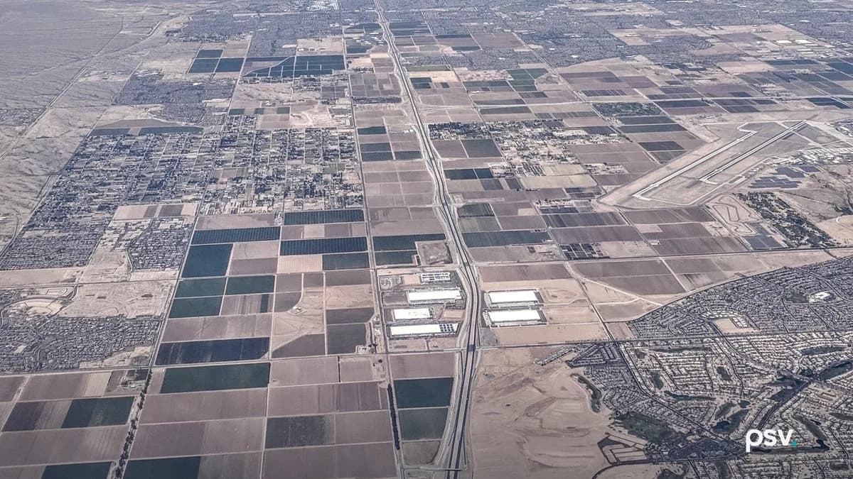 An aerial photograph looking north over the Phoenix West Valley in daylight, with Arizona State Route 303 running up the center as a divided highway through a grid of brown and green farm fields, a cluster of large white-roofed industrial buildings beside the freeway, subdivisions of houses at the right and left edges, the runways of Luke Air Force Base in the right background, and hazy desert mountains under a blue sky with thin clouds.