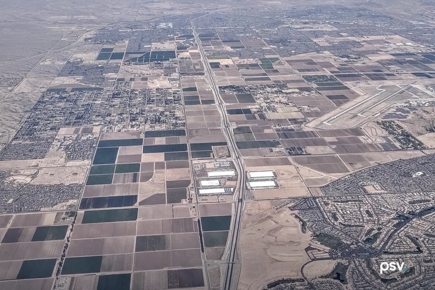 An aerial photograph looking north over the Phoenix West Valley in daylight, with Arizona State Route 303 running up the center as a divided highway through a grid of brown and green farm fields, a cluster of large white-roofed industrial buildings beside the freeway, subdivisions of houses at the right and left edges, the runways of Luke Air Force Base in the right background, and hazy desert mountains under a blue sky with thin clouds. An aerial photograph looking north over the Phoenix West Valley in daylight, with Arizona State Route 303 running up the center as a divided highway through a grid of brown and green farm fields, a cluster of large white-roofed industrial buildings beside the freeway, subdivisions of houses at the right and left edges, the runways of Luke Air Force Base in the right background, and hazy desert mountains under a blue sky with thin clouds.