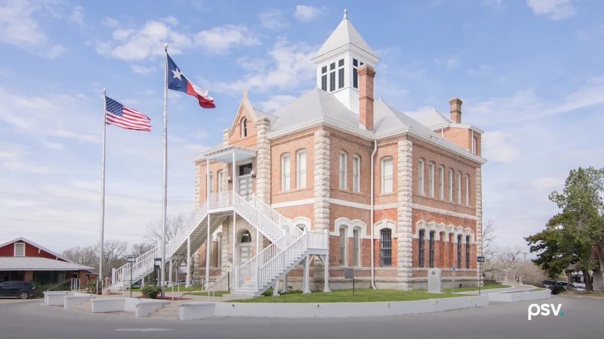 A daylight photograph of the Grimes County Courthouse in Anderson, Texas, seen from its corner across a curving paved street: a two-story red brick building with pale rusticated stone quoins, tall arched windows, barred windows on the ground floor at right, a grey hipped roof with a white square cupola and brick chimneys, and a white wooden double staircase climbing to a second-floor entrance porch. A United States flag and a Texas flag fly from two tall poles at left beside a low red building with a metal roof, a leafy tree stands at right, and a blue sky with scattered white clouds fills the upper half of the frame.
