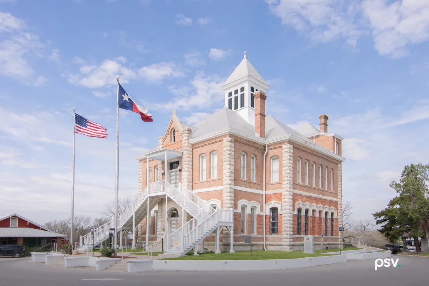 A daylight photograph of the Grimes County Courthouse in Anderson, Texas, seen from its corner across a curving paved street: a two-story red brick building with pale rusticated stone quoins, tall arched windows, barred windows on the ground floor at right, a grey hipped roof with a white square cupola and brick chimneys, and a white wooden double staircase climbing to a second-floor entrance porch. A United States flag and a Texas flag fly from two tall poles at left beside a low red building with a metal roof, a leafy tree stands at right, and a blue sky with scattered white clouds fills the upper half of the frame. A daylight photograph of the Grimes County Courthouse in Anderson, Texas, seen from its corner across a curving paved street: a two-story red brick building with pale rusticated stone quoins, tall arched windows, barred windows on the ground floor at right, a grey hipped roof with a white square cupola and brick chimneys, and a white wooden double staircase climbing to a second-floor entrance porch. A United States flag and a Texas flag fly from two tall poles at left beside a low red building with a metal roof, a leafy tree stands at right, and a blue sky with scattered white clouds fills the upper half of the frame.