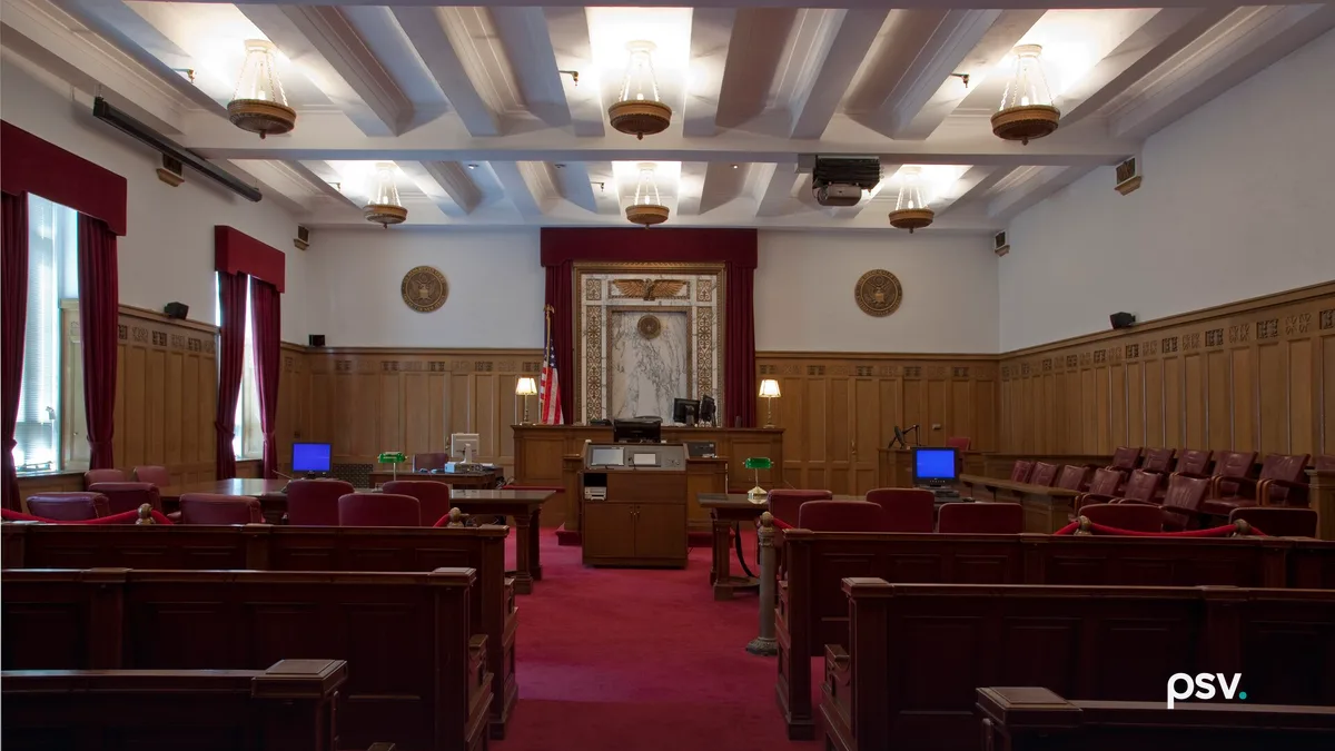 A photograph of an empty federal courtroom with dark wooden spectator benches and a red carpeted center aisle leading to a raised wooden judge’s bench, framed by a marble panel topped with a carved gilt eagle and red curtains, an American flag at the left, two circular seals on white walls above oak wainscoting, counsel tables with computer monitors and green desk lamps, a jury box of red chairs at the right, and a white beamed ceiling with hanging brass light fixtures.