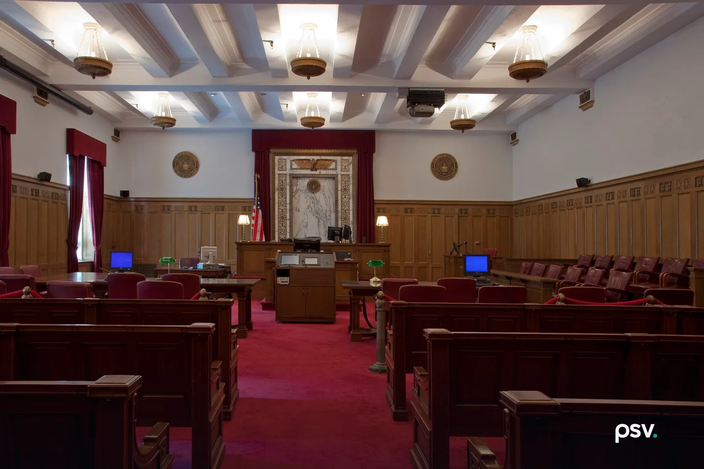 A photograph of an empty federal courtroom with dark wooden spectator benches and a red carpeted center aisle leading to a raised wooden judge’s bench, framed by a marble panel topped with a carved gilt eagle and red curtains, an American flag at the left, two circular seals on white walls above oak wainscoting, counsel tables with computer monitors and green desk lamps, a jury box of red chairs at the right, and a white beamed ceiling with hanging brass light fixtures. A photograph of an empty federal courtroom with dark wooden spectator benches and a red carpeted center aisle leading to a raised wooden judge’s bench, framed by a marble panel topped with a carved gilt eagle and red curtains, an American flag at the left, two circular seals on white walls above oak wainscoting, counsel tables with computer monitors and green desk lamps, a jury box of red chairs at the right, and a white beamed ceiling with hanging brass light fixtures.