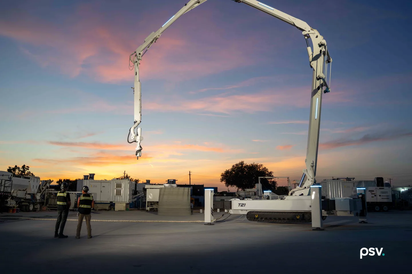 A white tracked robotic printer marked T21 at dusk, its long jointed boom arching high over an open concrete yard with a print nozzle hanging from the far end, a short grey layered printed wall section behind it, two workers in hard hats and high-visibility vests watching from the left, and equipment containers and a large tree against an orange and pink sunset sky. A white tracked robotic printer marked T21 at dusk, its long jointed boom arching high over an open concrete yard with a print nozzle hanging from the far end, a short grey layered printed wall section behind it, two workers in hard hats and high-visibility vests watching from the left, and equipment containers and a large tree against an orange and pink sunset sky.