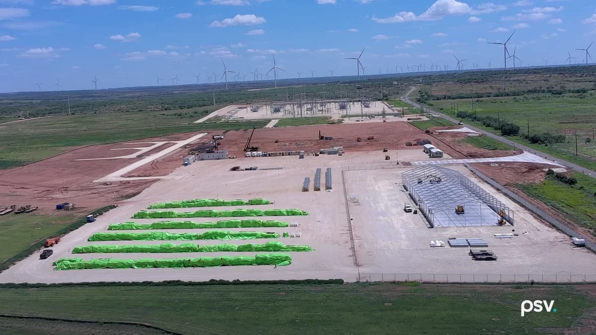 A daylight aerial view of IREN’s Sweetwater, Texas data center site under construction: a large graded pad of red earth ringed by security fence, a steel building frame going up on the right, rows of long green-wrapped equipment bundles staged on the left, site trailers and cranes in the middle distance, an electrical substation with steel lattice structures beyond the pad, and dozens of wind turbines spread across the flat horizon under a blue sky.