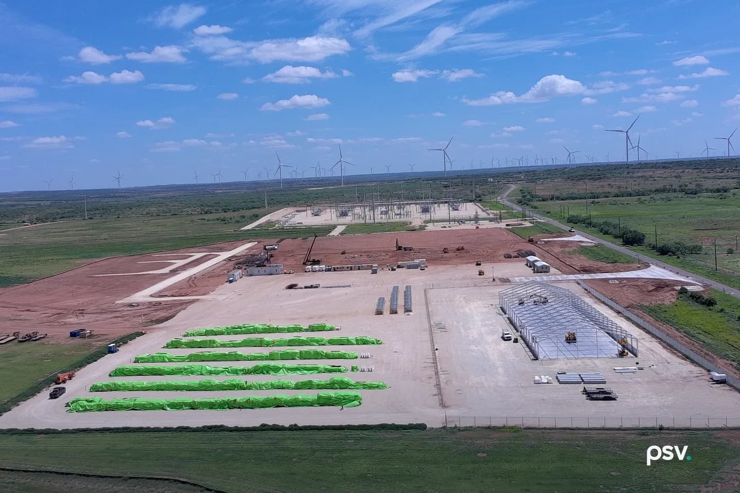 A daylight aerial view of IREN’s Sweetwater, Texas data center site under construction: a large graded pad of red earth ringed by security fence, a steel building frame going up on the right, rows of long green-wrapped equipment bundles staged on the left, site trailers and cranes in the middle distance, an electrical substation with steel lattice structures beyond the pad, and dozens of wind turbines spread across the flat horizon under a blue sky. A daylight aerial view of IREN’s Sweetwater, Texas data center site under construction: a large graded pad of red earth ringed by security fence, a steel building frame going up on the right, rows of long green-wrapped equipment bundles staged on the left, site trailers and cranes in the middle distance, an electrical substation with steel lattice structures beyond the pad, and dozens of wind turbines spread across the flat horizon under a blue sky.