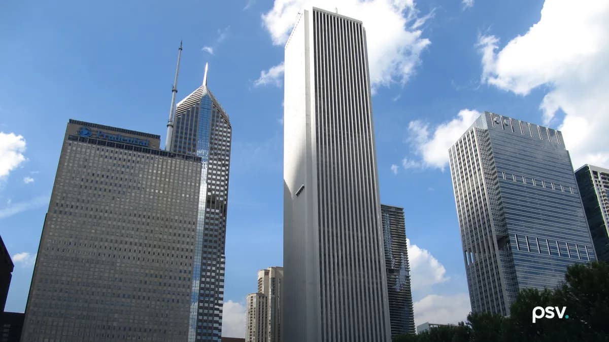 A daylight photograph looking up at Chicago’s Aon Center, a tall white tower with narrow vertical stripes, standing between the Prudential Plaza building with its rooftop sign and the spired Two Prudential Plaza at the left and a glass office tower at the right, under a blue sky with white clouds, with a row of green trees and park banners along the bottom.
