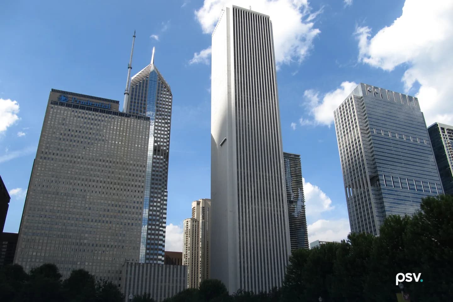 Looking up from street level at the tall, white, vertically ribbed Aon Center in the center of the frame, flanked by the Prudential Plaza towers on the left and a blue-glass office tower on the right, under a blue sky with scattered clouds. Looking up from street level at the tall, white, vertically ribbed Aon Center in the center of the frame, flanked by the Prudential Plaza towers on the left and a blue-glass office tower on the right, under a blue sky with scattered clouds.