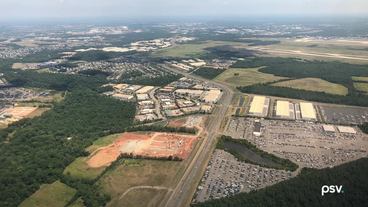 A daylight aerial photograph looking east from an airplane over Loudoun County, Virginia: a four-lane divided road runs from the bottom center toward the horizon, a cleared orange-red construction pad and a cluster of low white-roofed commercial and industrial buildings sit to its left, three long white-roofed warehouse buildings and large parking lots filled with rows of cars lie to its right beside a small pond, dense green woods fill the lower corners, and the runways of Washington Dulles International Airport stretch across the upper right under a hazy blue sky with a few white clouds.