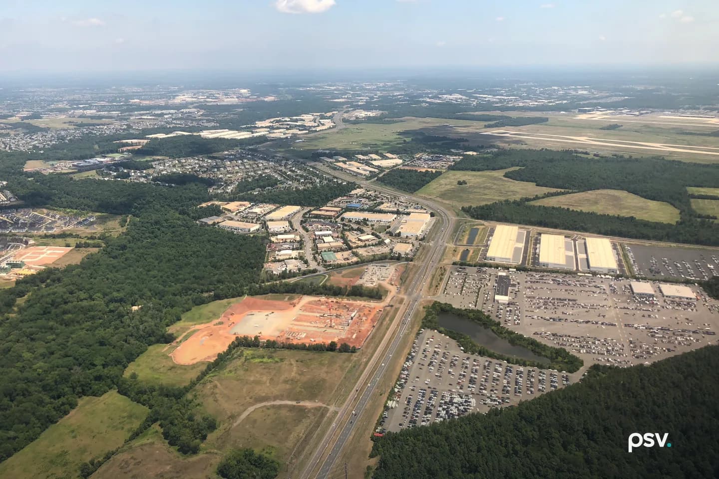 A daylight aerial photograph looking east from an airplane over Loudoun County, Virginia: a four-lane divided road runs from the bottom center toward the horizon, a cleared orange-red construction pad and a cluster of low white-roofed commercial and industrial buildings sit to its left, three long white-roofed warehouse buildings and large parking lots filled with rows of cars lie to its right beside a small pond, dense green woods fill the lower corners, and the runways of Washington Dulles International Airport stretch across the upper right under a hazy blue sky with a few white clouds. A daylight aerial photograph looking east from an airplane over Loudoun County, Virginia: a four-lane divided road runs from the bottom center toward the horizon, a cleared orange-red construction pad and a cluster of low white-roofed commercial and industrial buildings sit to its left, three long white-roofed warehouse buildings and large parking lots filled with rows of cars lie to its right beside a small pond, dense green woods fill the lower corners, and the runways of Washington Dulles International Airport stretch across the upper right under a hazy blue sky with a few white clouds.