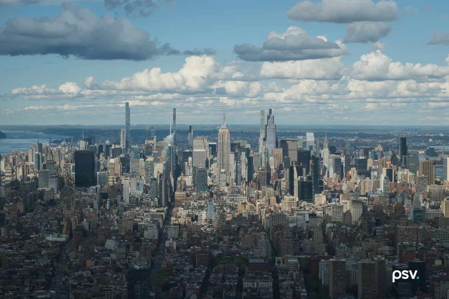 A daylight aerial photograph looking north over Manhattan from the One World Observatory: a dense low-rise field of brick and stone rooftops and water towers fills the foreground, stepping up through mid-block towers into the Midtown skyline across the middle of the frame, with the Empire State Building at center, slender pale supertall towers rising at left and right, the Chrysler Building’s stepped crown at right, the Hudson River and the New Jersey shoreline visible at far left, and a bright blue sky broken by scattered white cumulus above a flat horizon. A daylight aerial photograph looking north over Manhattan from the One World Observatory: a dense low-rise field of brick and stone rooftops and water towers fills the foreground, stepping up through mid-block towers into the Midtown skyline across the middle of the frame, with the Empire State Building at center, slender pale supertall towers rising at left and right, the Chrysler Building’s stepped crown at right, the Hudson River and the New Jersey shoreline visible at far left, and a bright blue sky broken by scattered white cumulus above a flat horizon.