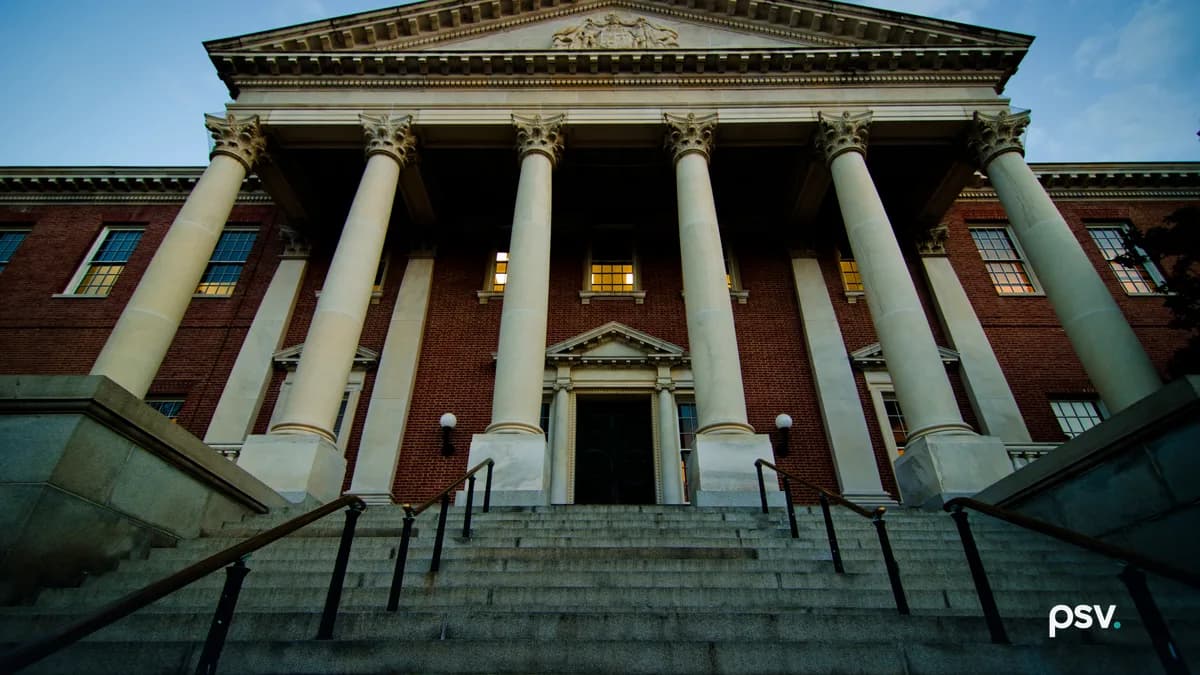 A low-angle photograph at dusk of the front of the Maryland State House in Annapolis, looking up a wide flight of granite steps divided by black metal handrails to a portico of six tall cream Corinthian columns under a carved pediment bearing the state seal, set against a red brick facade with lit multi-pane windows and a dark central doorway flanked by two globe lamps, beneath a blue sky with thin cloud.