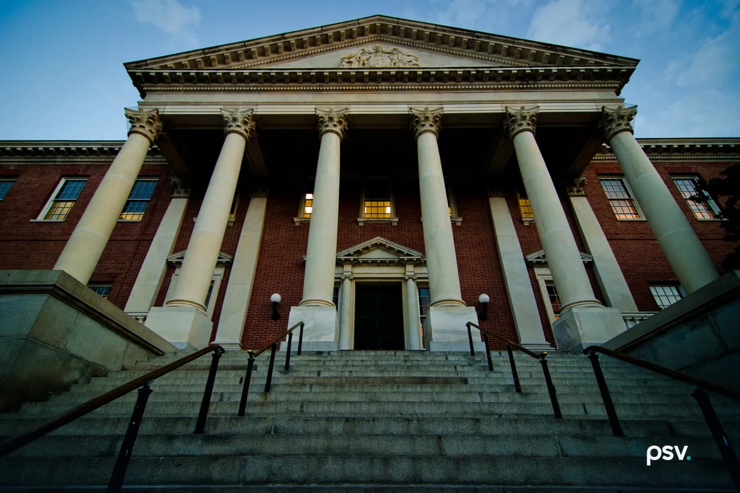 A low-angle photograph at dusk of the front of the Maryland State House in Annapolis, looking up a wide flight of granite steps divided by black metal handrails to a portico of six tall cream Corinthian columns under a carved pediment bearing the state seal, set against a red brick facade with lit multi-pane windows and a dark central doorway flanked by two globe lamps, beneath a blue sky with thin cloud. A low-angle photograph at dusk of the front of the Maryland State House in Annapolis, looking up a wide flight of granite steps divided by black metal handrails to a portico of six tall cream Corinthian columns under a carved pediment bearing the state seal, set against a red brick facade with lit multi-pane windows and a dark central doorway flanked by two globe lamps, beneath a blue sky with thin cloud.