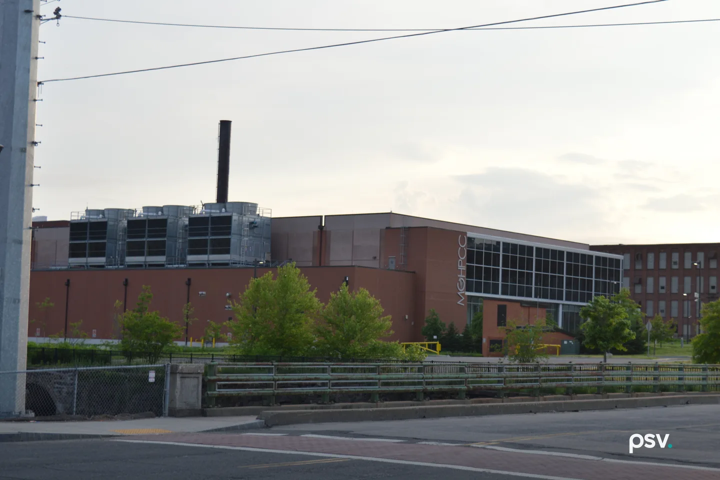 A photograph taken at dusk from across a paved street and a green steel bridge railing, looking at a long, low red brick data center building in Holyoke, Massachusetts: three banks of grey rooftop cooling units and a tall black exhaust stack rise on the left half of the roof, a glass-walled wing with vertical MGHPCC lettering on its brick corner sits at right, young leafy trees and a black fence line the strip in front, an older multi-story brick mill building stands at far right, and a concrete utility pole with overhead wires crosses a pale overcast sky at left. A photograph taken at dusk from across a paved street and a green steel bridge railing, looking at a long, low red brick data center building in Holyoke, Massachusetts: three banks of grey rooftop cooling units and a tall black exhaust stack rise on the left half of the roof, a glass-walled wing with vertical MGHPCC lettering on its brick corner sits at right, young leafy trees and a black fence line the strip in front, an older multi-story brick mill building stands at far right, and a concrete utility pole with overhead wires crosses a pale overcast sky at left.