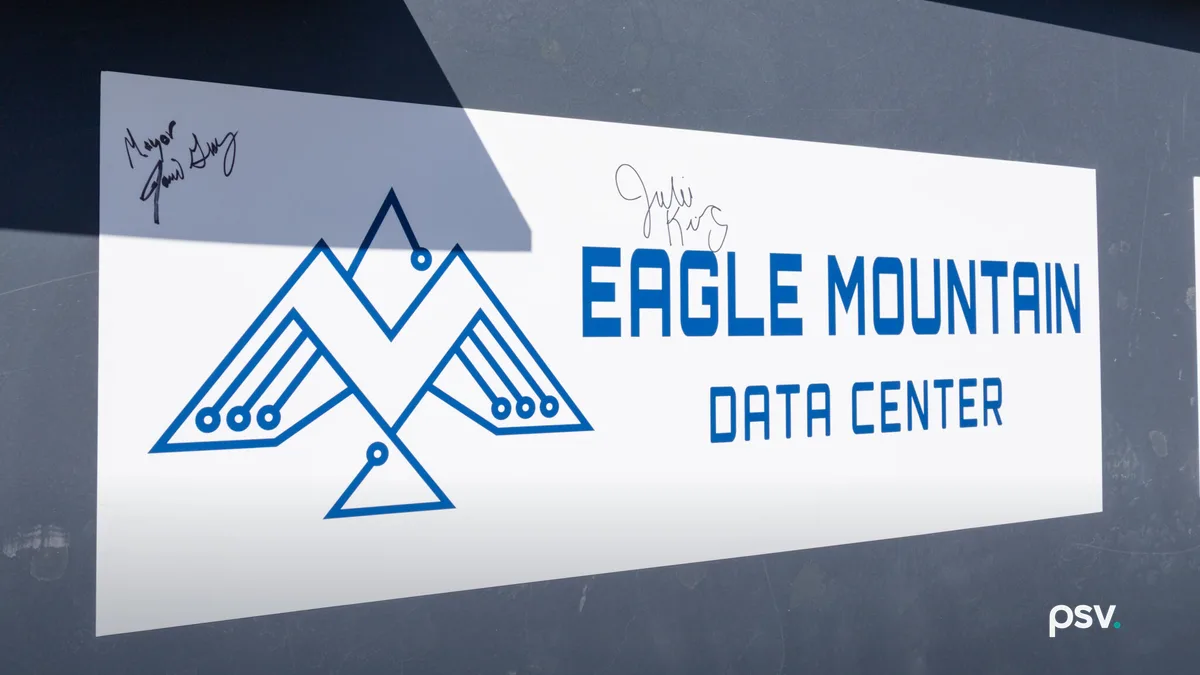 A close-up daylight photograph of a large white decal on a dark grey steel structural beam at a construction site, reading EAGLE MOUNTAIN DATA CENTER in blue block letters beside a blue line-drawn eagle formed from circuit traces and mountain peaks, with two handwritten signatures in black marker above it and a strip of blue sky at the upper right.