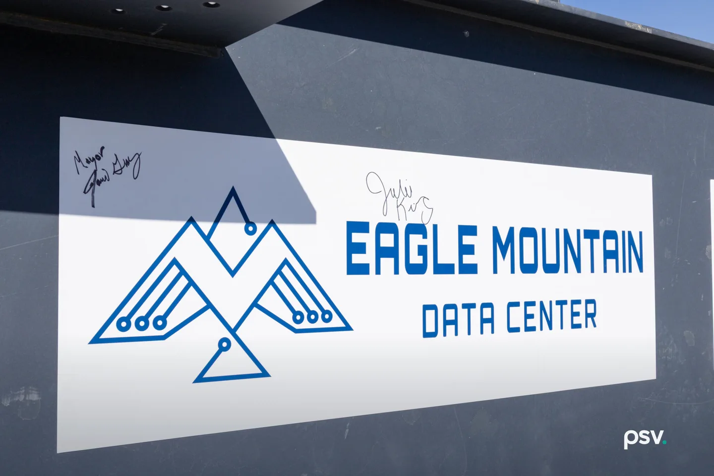 A close-up daylight photograph of a large white decal on a dark grey steel structural beam at a construction site, reading EAGLE MOUNTAIN DATA CENTER in blue block letters beside a blue line-drawn eagle formed from circuit traces and mountain peaks, with two handwritten signatures in black marker above it and a strip of blue sky at the upper right. A close-up daylight photograph of a large white decal on a dark grey steel structural beam at a construction site, reading EAGLE MOUNTAIN DATA CENTER in blue block letters beside a blue line-drawn eagle formed from circuit traces and mountain peaks, with two handwritten signatures in black marker above it and a strip of blue sky at the upper right.