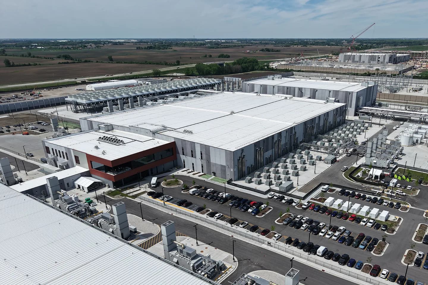 Aerial view of a completed Microsoft datacenter campus on flat Wisconsin farmland: long white-roofed data halls with rows of cooling units and electrical gear alongside, a full staff parking lot in the foreground, and a second facility under construction with a red crane on the horizon Aerial view of a completed Microsoft datacenter campus on flat Wisconsin farmland: long white-roofed data halls with rows of cooling units and electrical gear alongside, a full staff parking lot in the foreground, and a second facility under construction with a red crane on the horizon