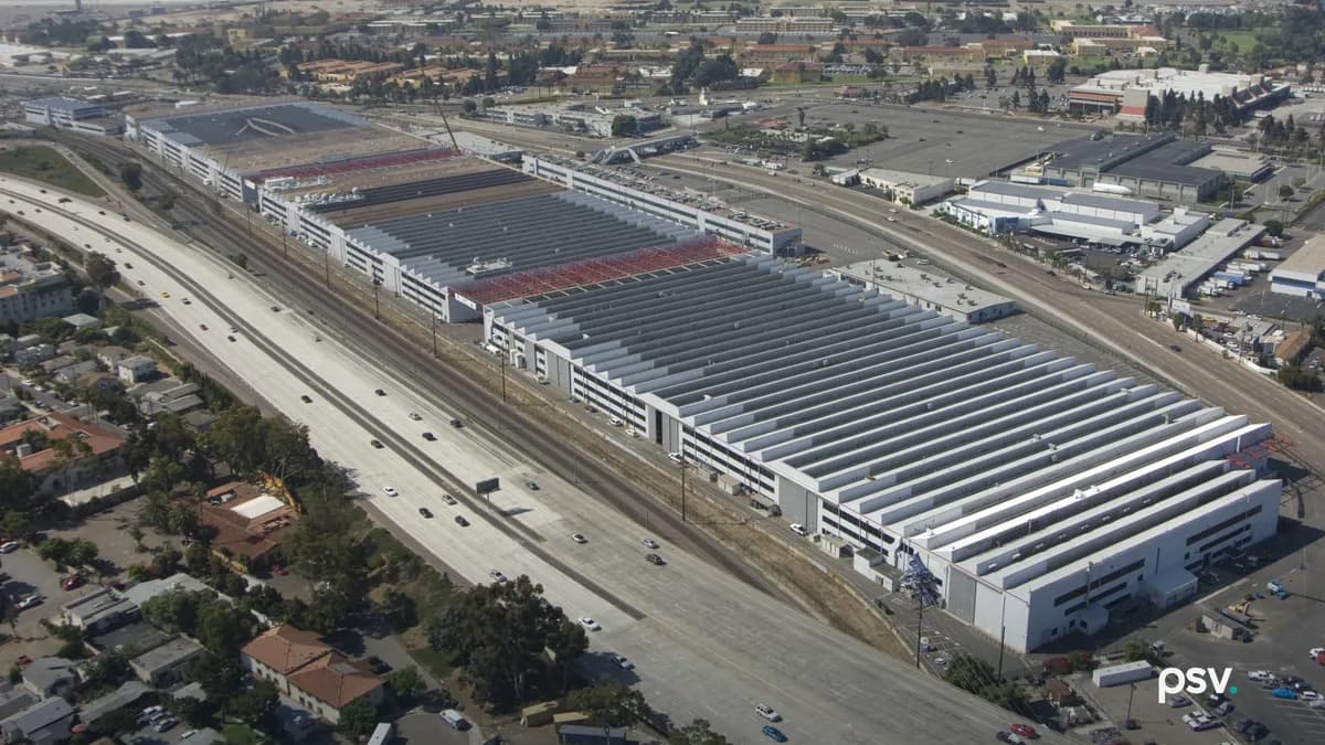An aerial view of the Navy’s Old Town Campus in San Diego: a row of long, low white industrial buildings with grey and red roofs running beside a multi-lane freeway and rail tracks, a neighborhood of houses and trees in the foreground, and an airport runway and San Diego Bay under a hazy sky in the distance.