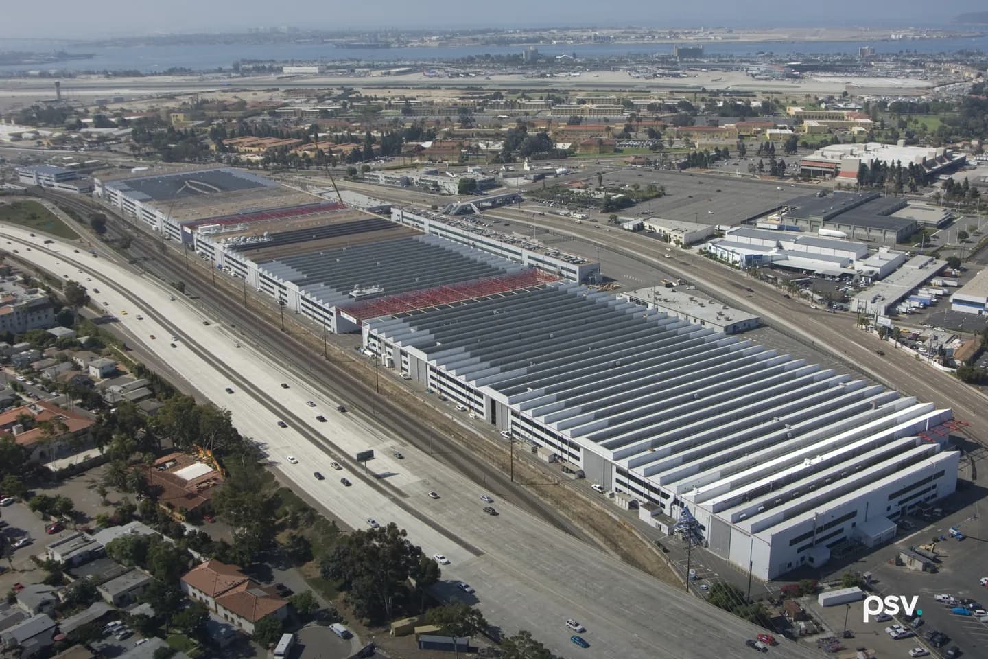 An aerial view of the Navy’s Old Town Campus in San Diego: a row of long, low white industrial buildings with grey and red roofs running beside a multi-lane freeway and rail tracks, a neighborhood of houses and trees in the foreground, and an airport runway and San Diego Bay under a hazy sky in the distance. An aerial view of the Navy’s Old Town Campus in San Diego: a row of long, low white industrial buildings with grey and red roofs running beside a multi-lane freeway and rail tracks, a neighborhood of houses and trees in the foreground, and an airport runway and San Diego Bay under a hazy sky in the distance.
