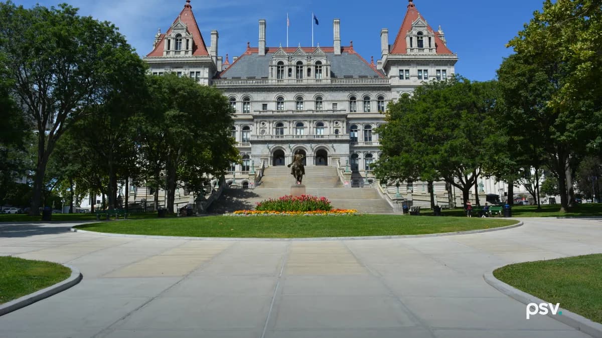 A daylight photograph of the New York State Capitol in Albany, a grey granite building with two red-roofed corner towers and a central mansard roof flying two flags, seen straight on across a curving concrete plaza and a lawn, with a bronze equestrian statue and a bed of red and yellow flowers at the foot of the wide front staircase and leafy green trees framing both sides under a blue sky.