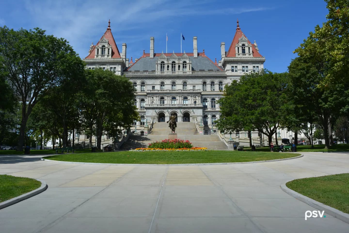 A daylight photograph of the New York State Capitol in Albany, a grey granite building with two red-roofed corner towers and a central mansard roof flying two flags, seen straight on across a curving concrete plaza and a lawn, with a bronze equestrian statue and a bed of red and yellow flowers at the foot of the wide front staircase and leafy green trees framing both sides under a blue sky. A daylight photograph of the New York State Capitol in Albany, a grey granite building with two red-roofed corner towers and a central mansard roof flying two flags, seen straight on across a curving concrete plaza and a lawn, with a bronze equestrian statue and a bed of red and yellow flowers at the foot of the wide front staircase and leafy green trees framing both sides under a blue sky.