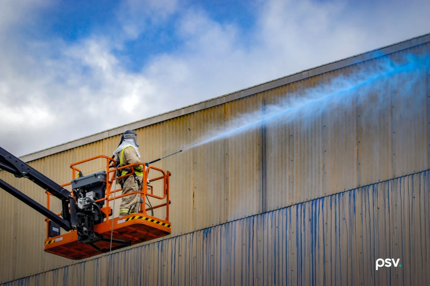 A worker in protective coveralls and a hood stands on the platform of an orange articulating boom lift, spraying a blue fixative mist across the tall corrugated metal wall of a decommissioned industrial process building, with blue streaks running down the lower panels and a partly cloudy sky behind. A worker in protective coveralls and a hood stands on the platform of an orange articulating boom lift, spraying a blue fixative mist across the tall corrugated metal wall of a decommissioned industrial process building, with blue streaks running down the lower panels and a partly cloudy sky behind.