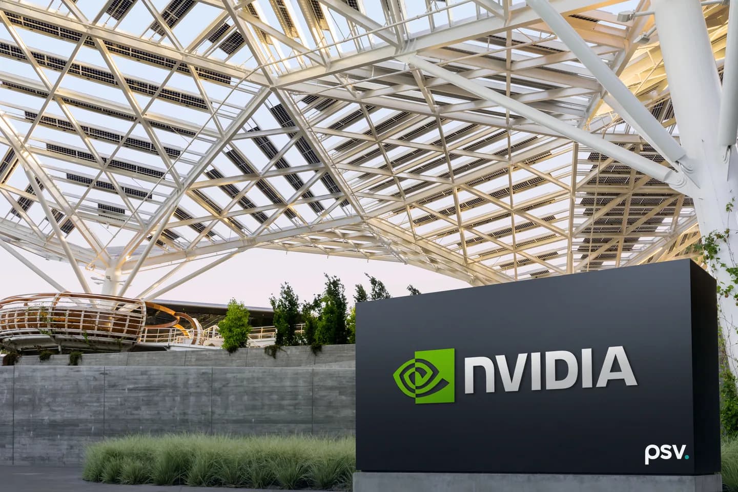 A dark monolith sign carrying the NVIDIA eye logo and wordmark standing on a concrete plinth in front of a board-formed concrete retaining wall, beneath a vast white triangulated steel canopy fitted with solar panels, with young trees, ornamental grasses and a curved walkway behind it at dusk A dark monolith sign carrying the NVIDIA eye logo and wordmark standing on a concrete plinth in front of a board-formed concrete retaining wall, beneath a vast white triangulated steel canopy fitted with solar panels, with young trees, ornamental grasses and a curved walkway behind it at dusk
