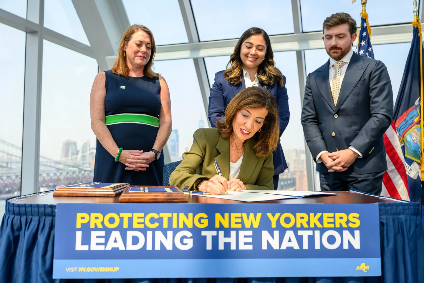 Governor Kathy Hochul signing Executive Order 62 at a table with a Protecting New Yorkers banner, with staff standing behind her Governor Kathy Hochul signing Executive Order 62 at a table with a Protecting New Yorkers banner, with staff standing behind her
