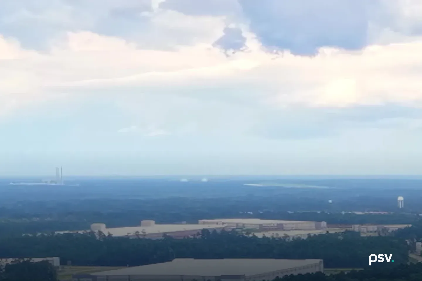Wide aerial view over Effingham County, Georgia on a cloudy day: long white distribution warehouses and storage tanks set among dense pine forest, with a water tower and a power plant stack on the flat horizon Wide aerial view over Effingham County, Georgia on a cloudy day: long white distribution warehouses and storage tanks set among dense pine forest, with a water tower and a power plant stack on the flat horizon