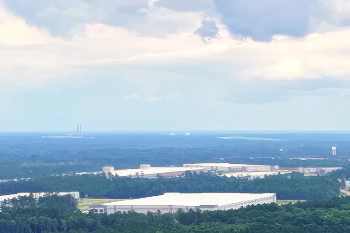 Wide aerial view over Effingham County, Georgia on a cloudy day: long white distribution warehouses and storage tanks set among dense pine forest, with a water tower and a power plant stack on the flat horizon Wide aerial view over Effingham County, Georgia on a cloudy day: long white distribution warehouses and storage tanks set among dense pine forest, with a water tower and a power plant stack on the flat horizon