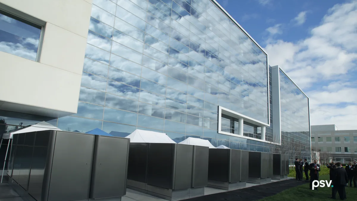A daylight photograph of a row of grey stainless steel fuel cell cabinets on a concrete pad beside a modern office building clad in a mirrored glass curtain wall reflecting white clouds and blue sky, with a cream stone wing at the left, white event tents behind the cabinets, a strip of lawn and a black rubber walkway in the foreground, and a small group of people in suits standing on the grass at the right.