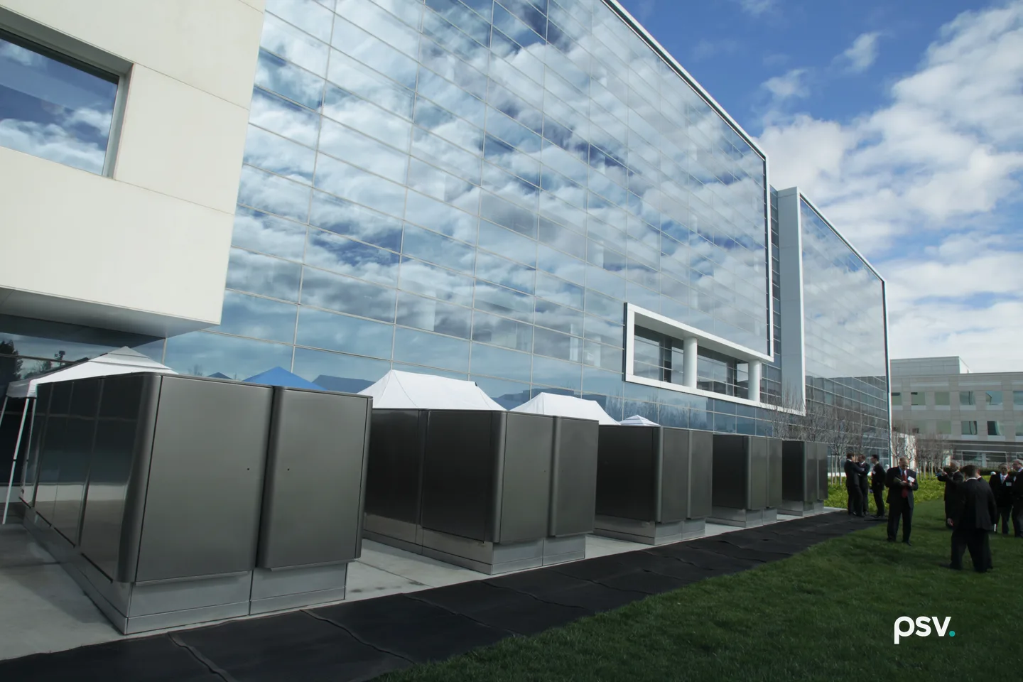 A daylight photograph of a row of grey stainless steel fuel cell cabinets on a concrete pad beside a modern office building clad in a mirrored glass curtain wall reflecting white clouds and blue sky, with a cream stone wing at the left, white event tents behind the cabinets, a strip of lawn and a black rubber walkway in the foreground, and a small group of people in suits standing on the grass at the right. A daylight photograph of a row of grey stainless steel fuel cell cabinets on a concrete pad beside a modern office building clad in a mirrored glass curtain wall reflecting white clouds and blue sky, with a cream stone wing at the left, white event tents behind the cabinets, a strip of lawn and a black rubber walkway in the foreground, and a small group of people in suits standing on the grass at the right.