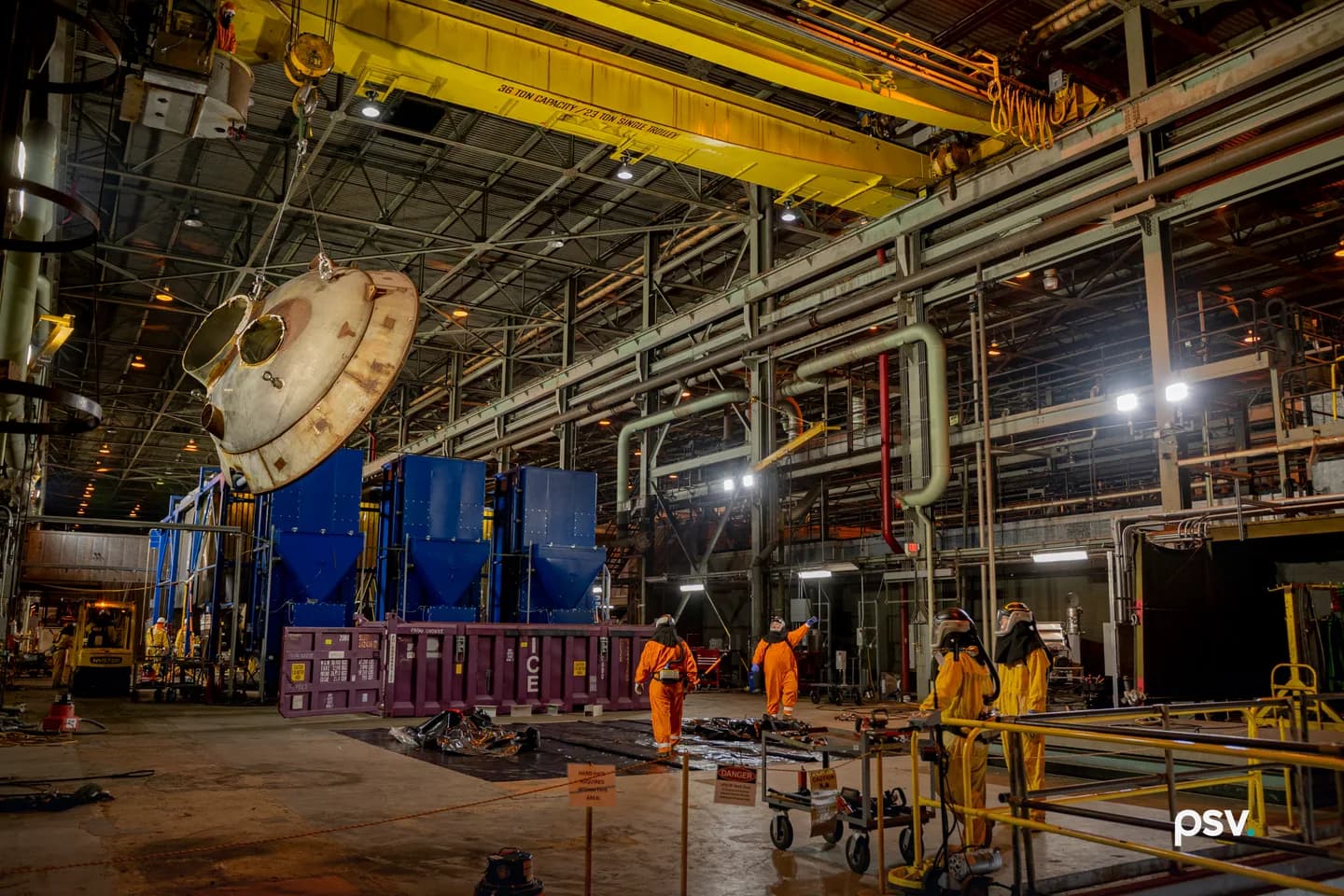 Interior of a vast industrial building at the Department of Energy’s Paducah Site: a yellow 36-ton overhead crane lifts a large corroded metal vessel head while workers in orange and yellow protective suits and respirators stand on the floor below beside a purple waste container and blue processing equipment Interior of a vast industrial building at the Department of Energy’s Paducah Site: a yellow 36-ton overhead crane lifts a large corroded metal vessel head while workers in orange and yellow protective suits and respirators stand on the floor below beside a purple waste container and blue processing equipment