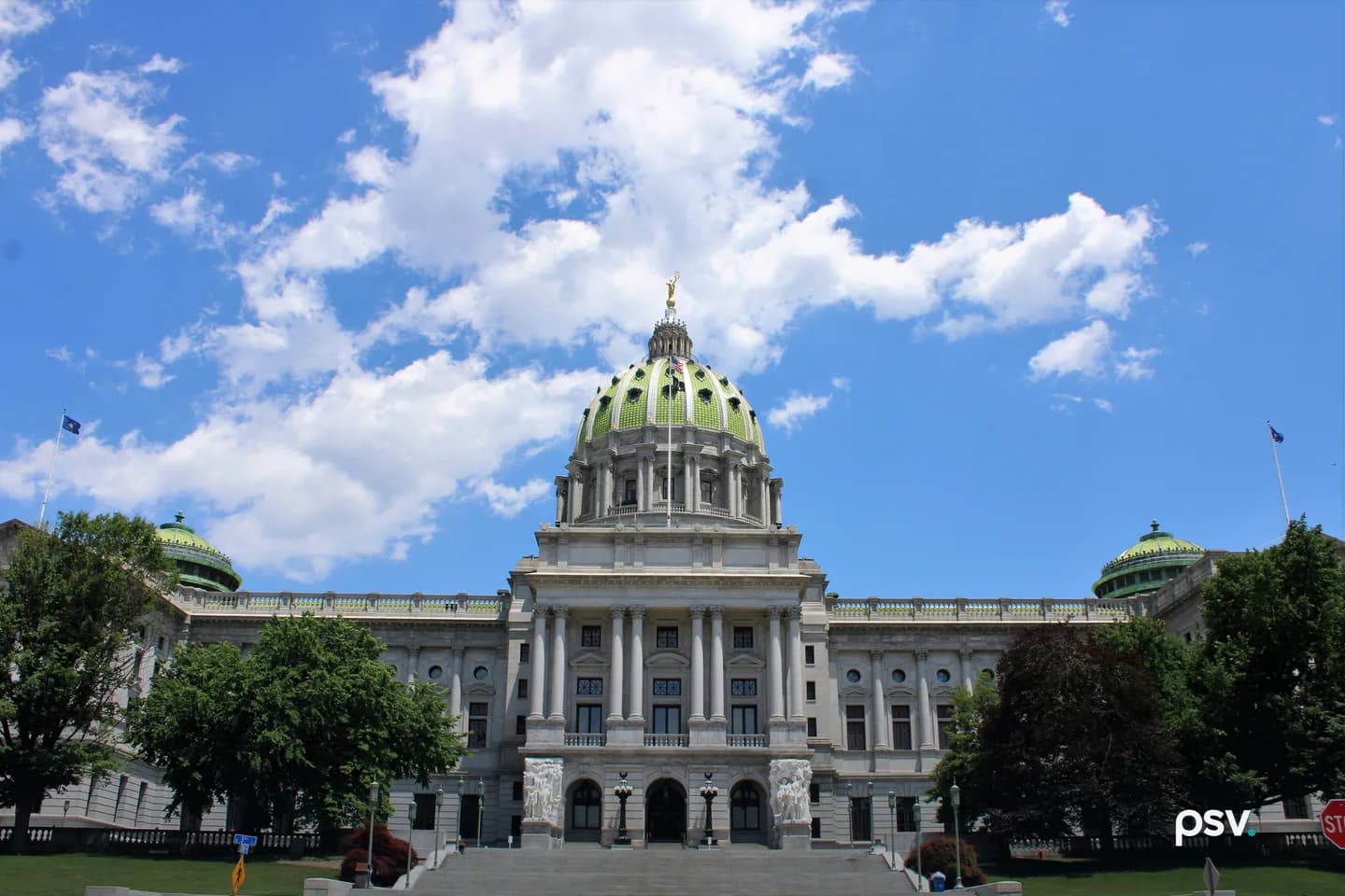 The Pennsylvania State Capitol in Harrisburg photographed from the front steps on a clear summer day: a grey granite Beaux-Arts building with a tall columned portico, a green tiled dome topped by a gilded statue, matching green domes over the wings, flanking trees, and scattered cumulus cloud overhead The Pennsylvania State Capitol in Harrisburg photographed from the front steps on a clear summer day: a grey granite Beaux-Arts building with a tall columned portico, a green tiled dome topped by a gilded statue, matching green domes over the wings, flanking trees, and scattered cumulus cloud overhead