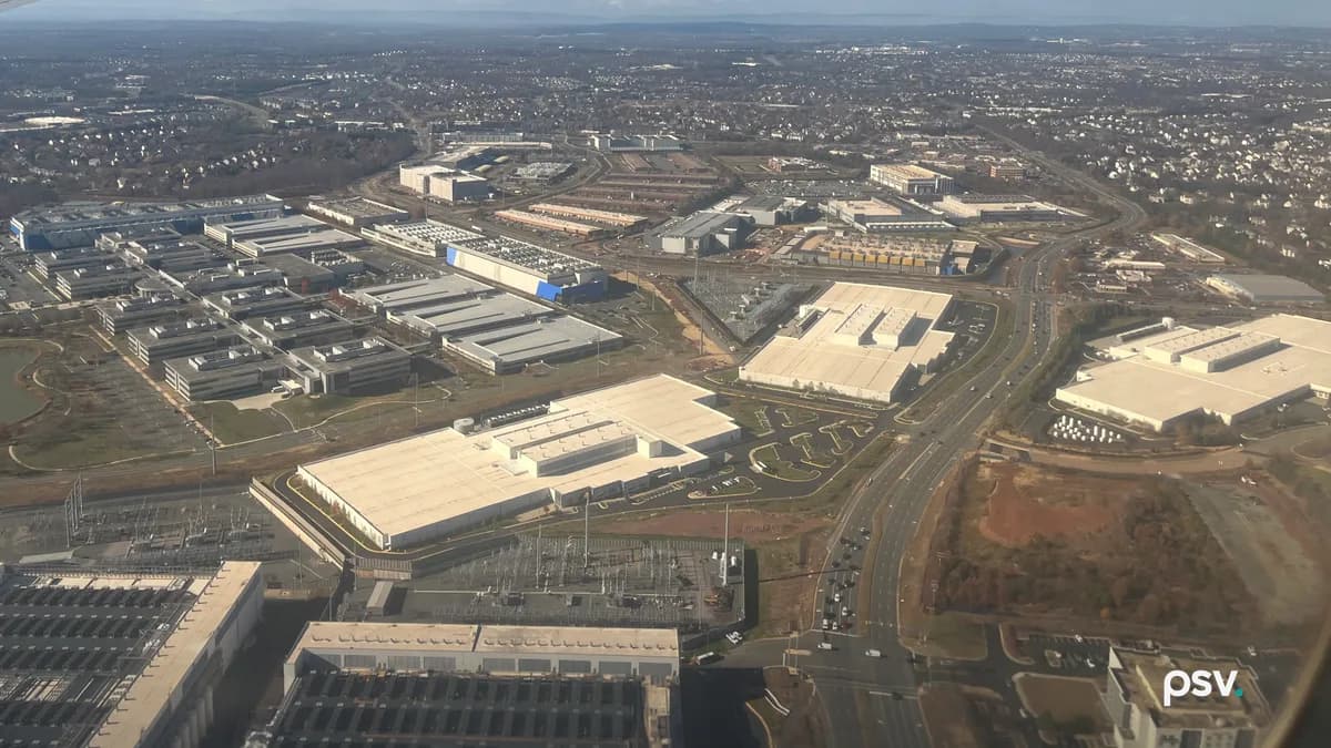 A daylight aerial photograph over Loudoun County, Virginia, looking across a dense field of low, flat-roofed data center buildings in pale grey and white, arranged in long parallel rows with rooftop cooling equipment, separated by wide access roads and parking aprons; two fenced electrical substations with lattice steel structures and busbars sit in the near foreground, a multi-lane divided highway runs diagonally through the frame at right past bare red-brown graded ground, and low suburban rooftops and tree cover stretch to a hazy ridgeline on the horizon.